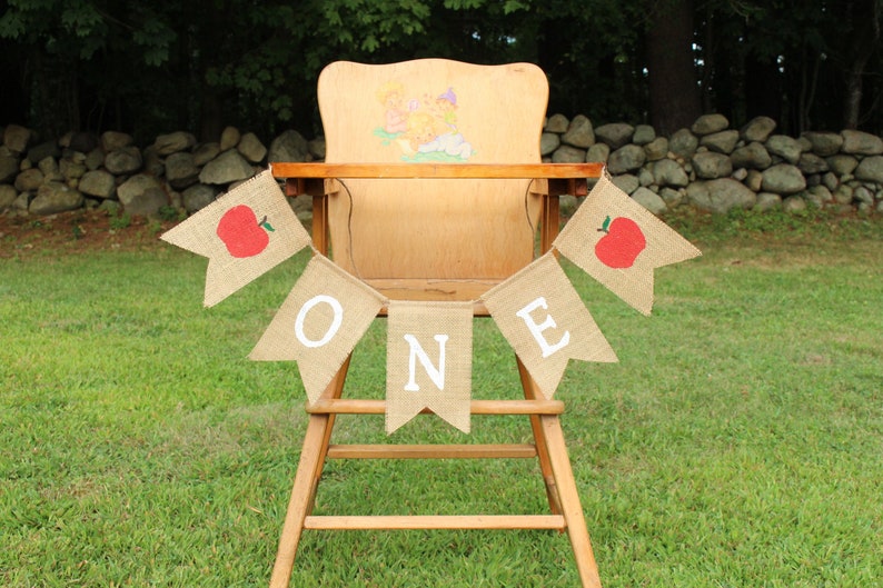 May include: A burlap banner with red apples and white letters spelling "ONE" hangs on a wooden high chair.