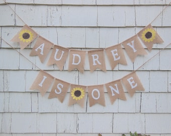 May include: A burlap banner with the text "AUDREY IS ONE" in white letters. Each pennant is tan with a sunflower design and strung on twine. The banner is displayed against a white, weathered wood background.