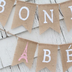 May include: A burlap banner with the words "Bonjour" and a pink Eiffel Tower graphic. The banner is strung on a white wooden surface.