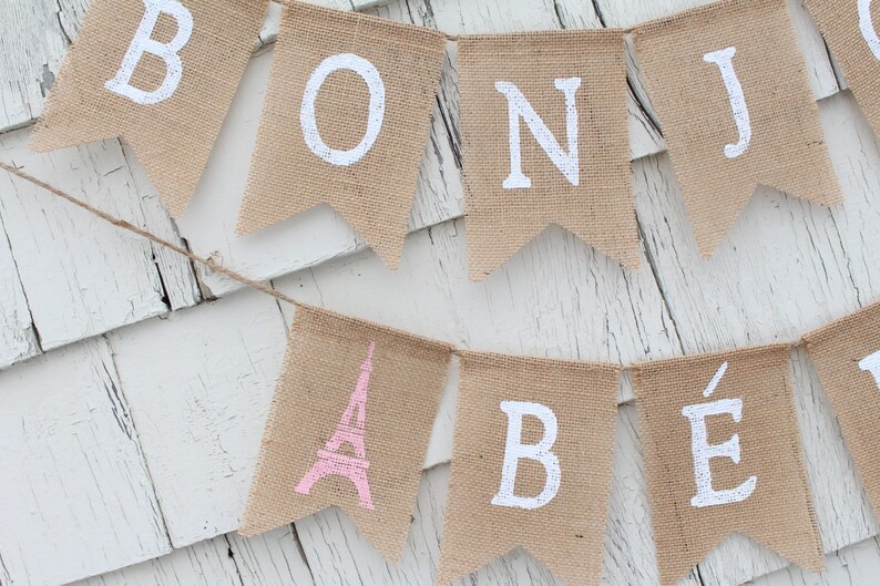 May include: A burlap banner with the words "Bonjour" and a pink Eiffel Tower graphic. The banner is strung on a white wooden surface.