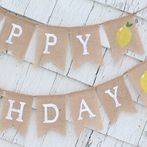 May include: A burlap banner with the words "Happy Birthday" in white paint. The banner is decorated with yellow lemons with green leaves.