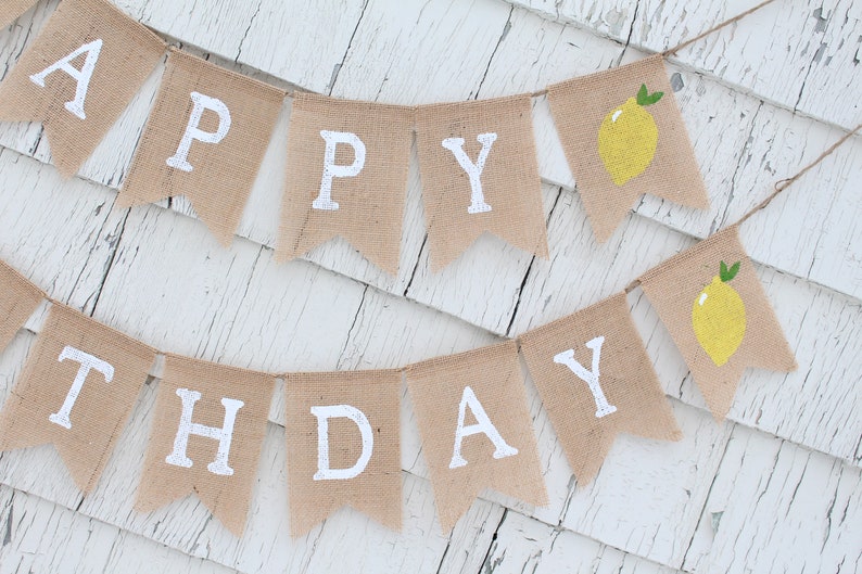 May include: A burlap banner with the words "Happy Birthday" in white paint. The banner is decorated with yellow lemons with green leaves.