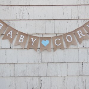May include: A burlap banner with the words "Baby Cory" in white letters. A light blue heart is between the words.