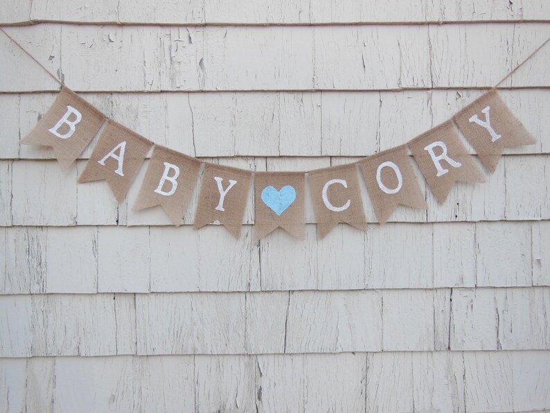 May include: A burlap banner with the words "Baby Cory" in white letters. A light blue heart is between the words.