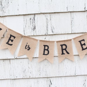 May include: A burlap banner with the text "LOVE is BREW" in black lettering. The banner is hanging on a white wooden wall.