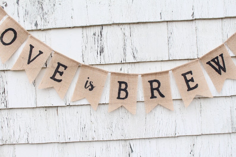 May include: A burlap banner with the text "LOVE is BREW" in black lettering. The banner is hanging on a white wooden wall.
