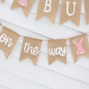 May include: A burlap banner with the words "It's a Bun on the Way" in white paint. The banner is decorated with pink bunny cutouts.