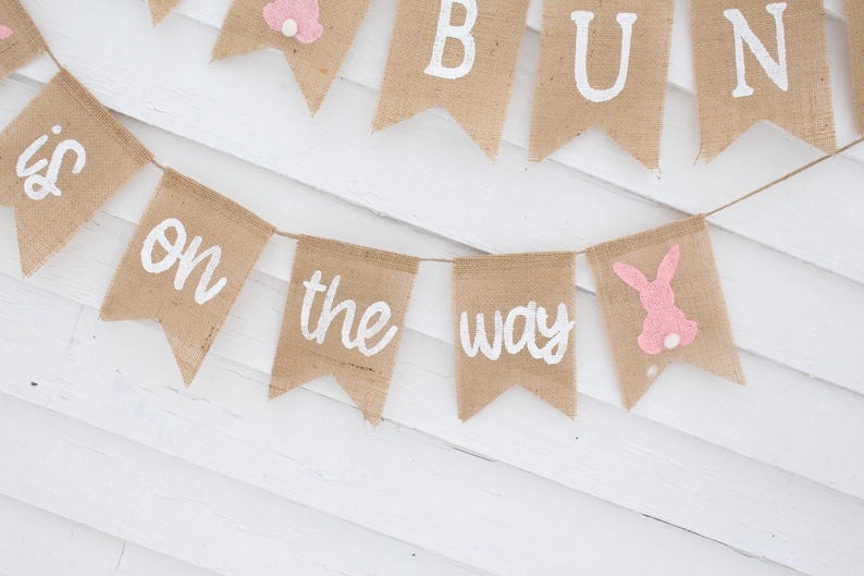 May include: A burlap banner with the words "It's a Bun on the Way" in white paint. The banner is decorated with pink bunny cutouts.