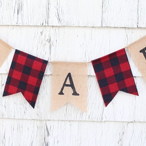 May include: A burlap banner with the letters "S A B" in black, with red and black buffalo plaid fabric flags between each letter.
