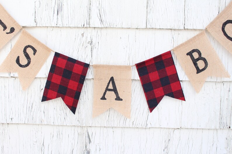 May include: A burlap banner with the letters "S A B" in black, with red and black buffalo plaid fabric flags between each letter.