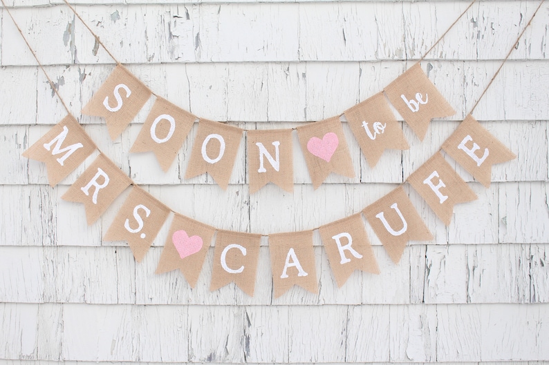 May include: A burlap banner with the text "Mr. & Mrs. Soon to be Carufe" in white lettering. The banner has pink hearts between the words.