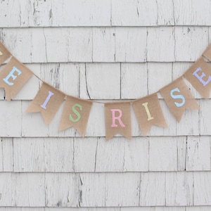 May include: A burlap banner with the words "He is Risen" in pastel colors. The banner is hanging on a white wooden wall.