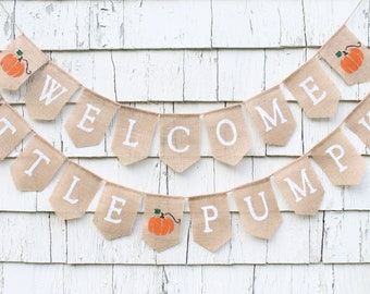 Könnte beinhalten: Ein Banner aus Jute mit den Worten "Welcome Little Pumpkin" in weißen Buchstaben. Das Banner ist mit kleinen orangefarbenen Kürbissen verziert.
