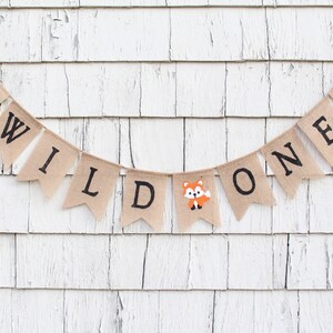 May include: A burlap banner with the words "WILD ONE" in black letters. The banner is decorated with small animal illustrations, including a raccoon, a fox, and a bear.