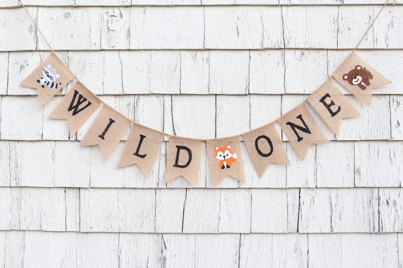 May include: A burlap banner with the words "WILD ONE" in black letters. The banner is decorated with small animal illustrations, including a raccoon, a fox, and a bear.