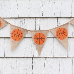 Puede incluir: Una pancarta de arpillera con balones de baloncesto naranjas impresos en cada bandera triangular. La pancarta est&aacute; colgada en una pared de madera blanca.