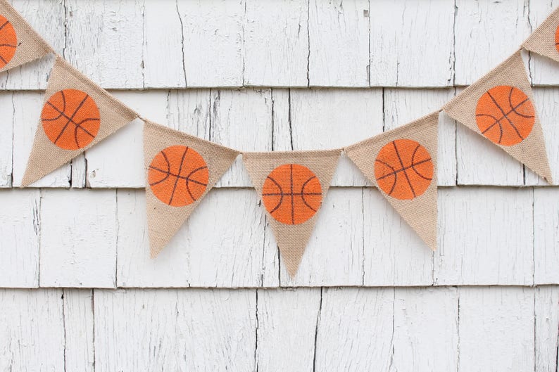 Puede incluir: Una pancarta de arpillera con balones de baloncesto naranjas impresos en cada bandera triangular. La pancarta est&aacute; colgada en una pared de madera blanca.