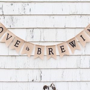 May include: A burlap banner with the text "LOVE is BREWING" in black lettering. The banner is strung up against a white wooden wall.