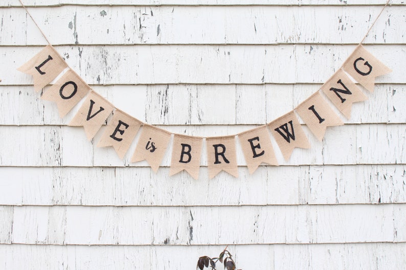 May include: A burlap banner with the text "LOVE is BREWING" in black lettering. The banner is strung up against a white wooden wall.