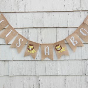May include: A burlap banner with the words "It's a Boy" in white lettering. The banner has small lion illustrations on each of the flags.