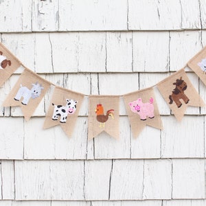 Puede incluir: Una pancarta de arpillera con ilustraciones de animales de granja. La pancarta presenta un perro, una cabra, una vaca, un gallo, un cerdo y un caballo. La pancarta est&aacute; colgada en un fondo de madera blanca.