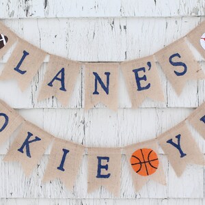 Pu&ograve; includere: Uno striscione di juta con il testo "LANE'S ROOKIE YEAR" in lettere blu. Lo striscione &egrave; decorato con un pallone da calcio, baseball e pallacanestro.