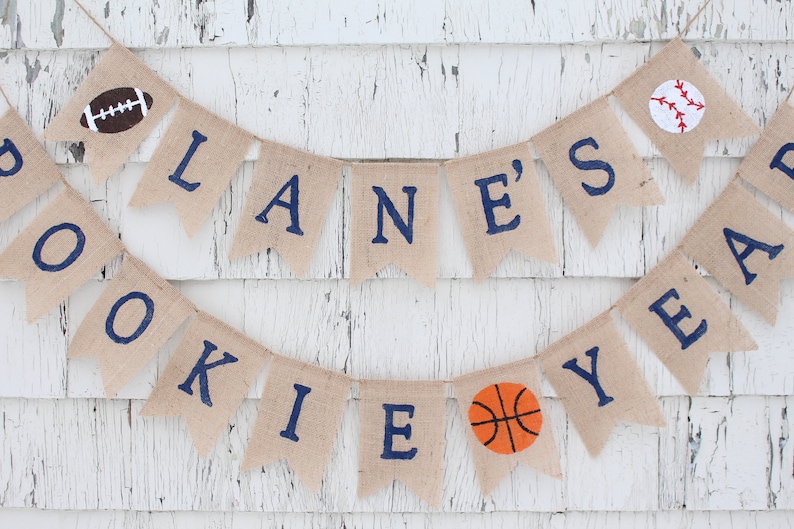 Pu&ograve; includere: Uno striscione di juta con il testo "LANE'S ROOKIE YEAR" in lettere blu. Lo striscione &egrave; decorato con un pallone da calcio, baseball e pallacanestro.