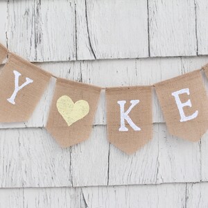 May include: A burlap banner with the words "BY" and "KEN" in white letters, separated by a yellow heart. The banner is hanging on a white wooden wall.