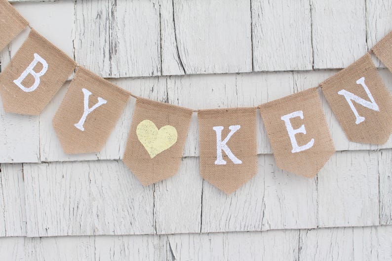 May include: A burlap banner with the words "BY" and "KEN" in white letters, separated by a yellow heart. The banner is hanging on a white wooden wall.