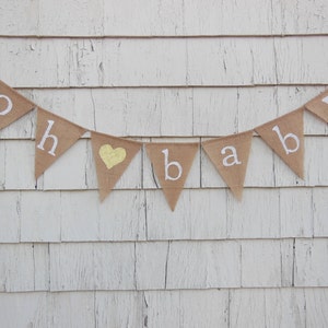 May include: A burlap banner with the words "Oh baby" in white letters and a gold heart. The banner is strung on a white wooden wall.
