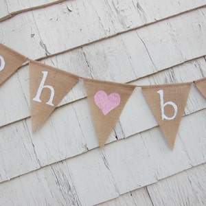 May include: A burlap banner with the words "Oh Baby" in white paint. A pink heart is in the middle of the banner.