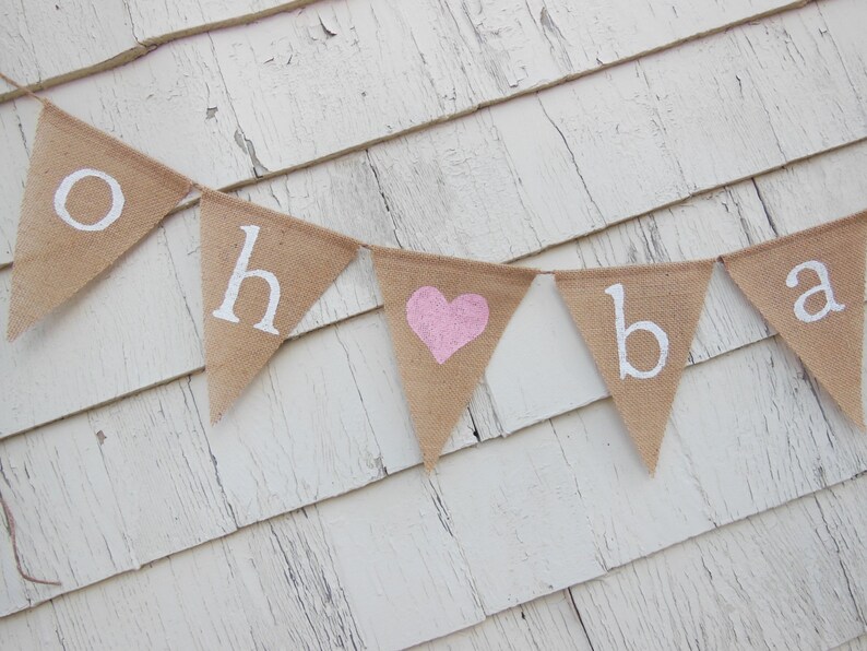 May include: A burlap banner with the words "Oh Baby" in white paint. A pink heart is in the middle of the banner.