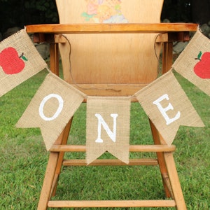 May include: A burlap banner with the word "ONE" in white letters and red apple accents hangs on a wooden high chair. The banner is perfect for a first birthday party.