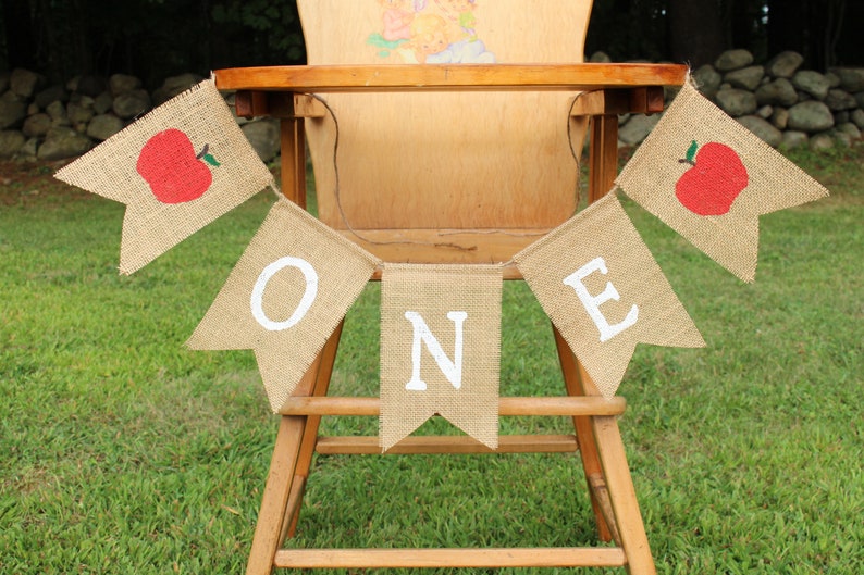 May include: A burlap banner with the word "ONE" in white letters and red apple accents hangs on a wooden high chair. The banner is perfect for a first birthday party.