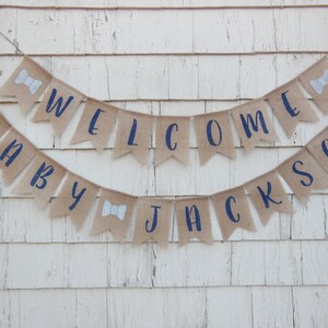 May include: A burlap banner with navy blue lettering that reads "Baby Jackson Welcome". The banner is decorated with white bowties.
