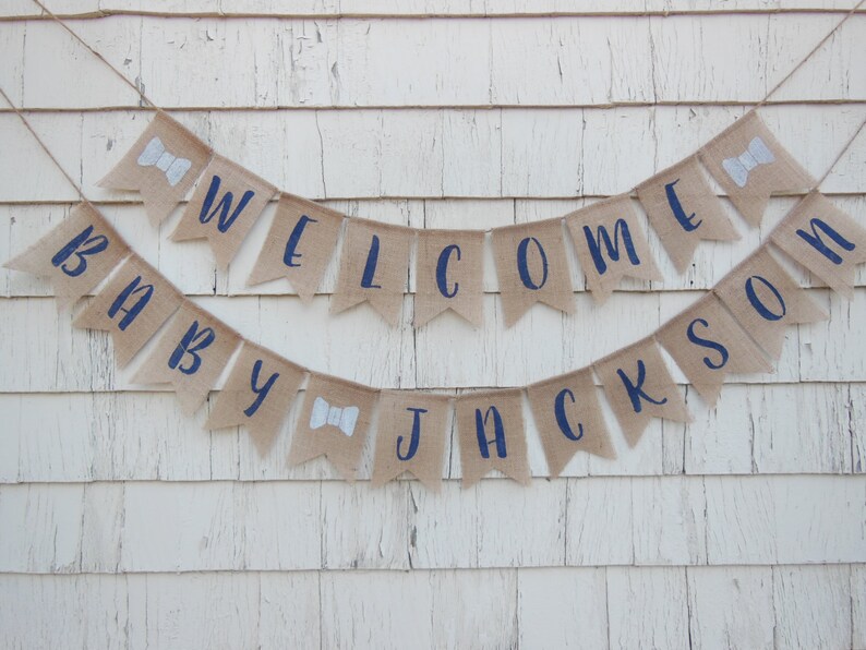 May include: A burlap banner with navy blue lettering that reads "Baby Jackson Welcome". The banner is decorated with white bowties.