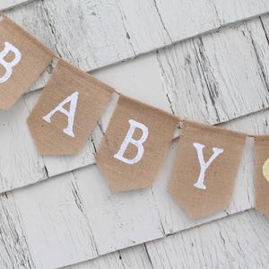 May include: A burlap banner with the word "BABY" in white letters and a yellow heart. The banner is hanging on a white wooden wall.