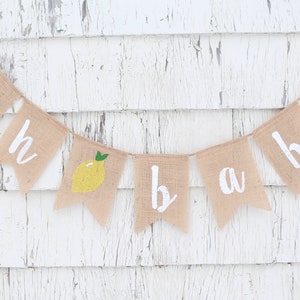 Peut inclure: Une banni&egrave;re en toile de jute avec les mots "oh baby" peints en blanc. Chaque lettre est sur un drapeau triangulaire s&eacute;par&eacute;. Les drapeaux sont d&eacute;cor&eacute;s de citrons jaunes avec des feuilles vertes.