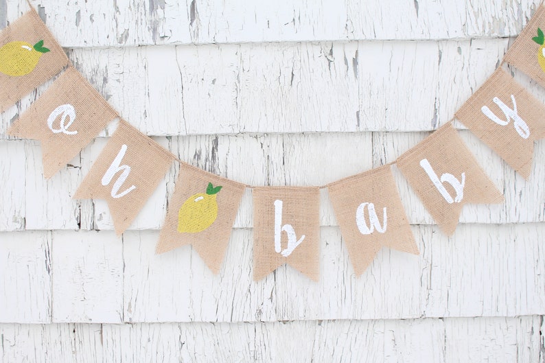Peut inclure: Une banni&egrave;re en toile de jute avec les mots "oh baby" peints en blanc. Chaque lettre est sur un drapeau triangulaire s&eacute;par&eacute;. Les drapeaux sont d&eacute;cor&eacute;s de citrons jaunes avec des feuilles vertes.