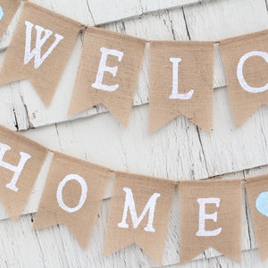 May include: A burlap banner with the words "WELCOME HOME" in white paint. The banner has two light blue hearts on either end.