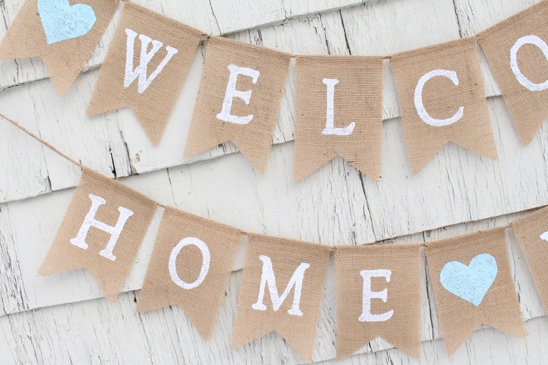 May include: A burlap banner with the words "WELCOME HOME" in white paint. The banner has two light blue hearts on either end.