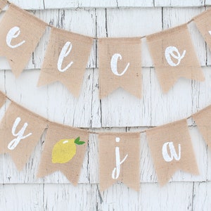 May include: A burlap banner with the words "Welcome to our July Jam" in white paint. A yellow lemon with green leaves is included in the banner.