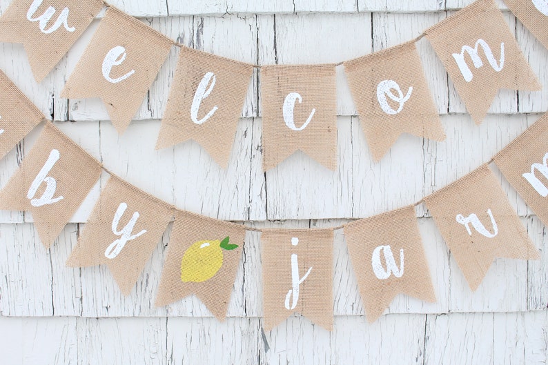 May include: A burlap banner with the words "Welcome to our July Jam" in white paint. A yellow lemon with green leaves is included in the banner.