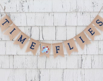May include: A burlap banner with the words "TIME FLIES" in navy blue lettering. A small blue aeroplane with red accents is between the words "TIME" and "FLIES". The banner is hanging on a white wooden wall.
