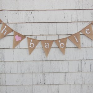 May include: A burlap banner with the words "Oh &hearts; Babies" in white lettering. The heart is pink.