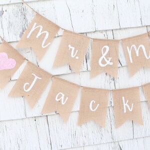 K&ouml;nnte beinhalten: Ein Banner aus Jute mit den Worten "Mr. & Mrs. Jack & Ky" in wei&szlig;er Farbe. Ein rosa Herz befindet sich auf der linken Seite des Banners.