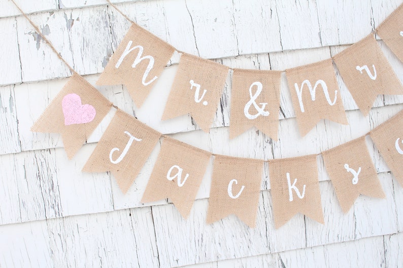 K&ouml;nnte beinhalten: Ein Banner aus Jute mit den Worten "Mr. & Mrs. Jack & Ky" in wei&szlig;er Farbe. Ein rosa Herz befindet sich auf der linken Seite des Banners.