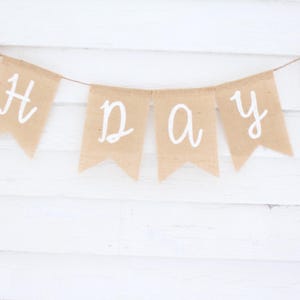 May include: A banner made of tan burlap flags strung together with white lettering that reads "CH DAY 2". The banner is hanging against a white wooden background.