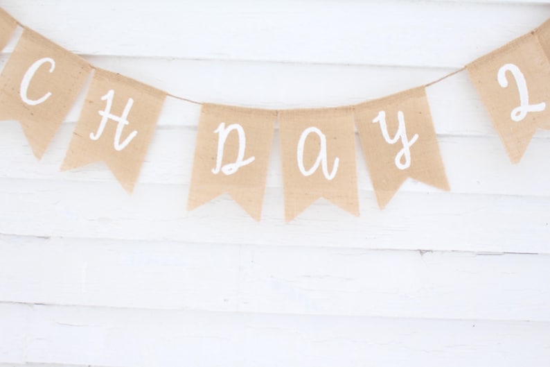 May include: A banner made of tan burlap flags strung together with white lettering that reads "CH DAY 2". The banner is hanging against a white wooden background.