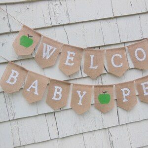 K&ouml;nnte beinhalten: Ein Wimpelbanner aus Jute mit den wei&szlig;en Gro&szlig;buchstaben "WELCOME BABY". Das Banner enth&auml;lt zwei gr&uuml;ne Apfelillustrationen. Es ist an einer d&uuml;nnen Schnur befestigt und h&auml;ngt vor einem wei&szlig;en, verwitterten Holzhintergrund.